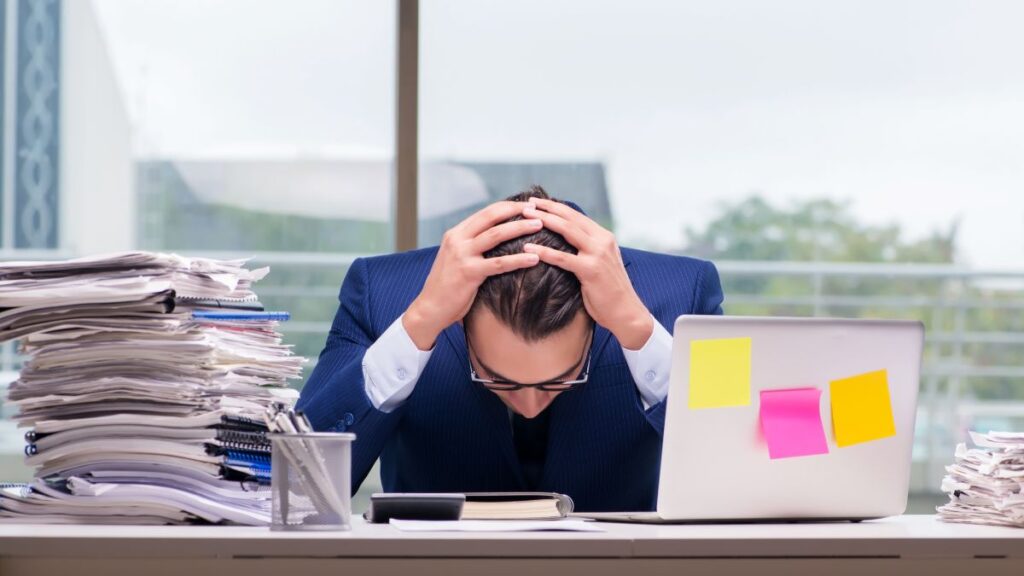 Man sitting with his head in his hands at his desk with work piled up