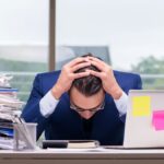 Man sitting with his head in his hands at his desk with work piled up