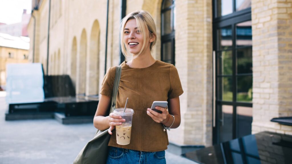 Woman walking down the street holding her phone and an iced coffee